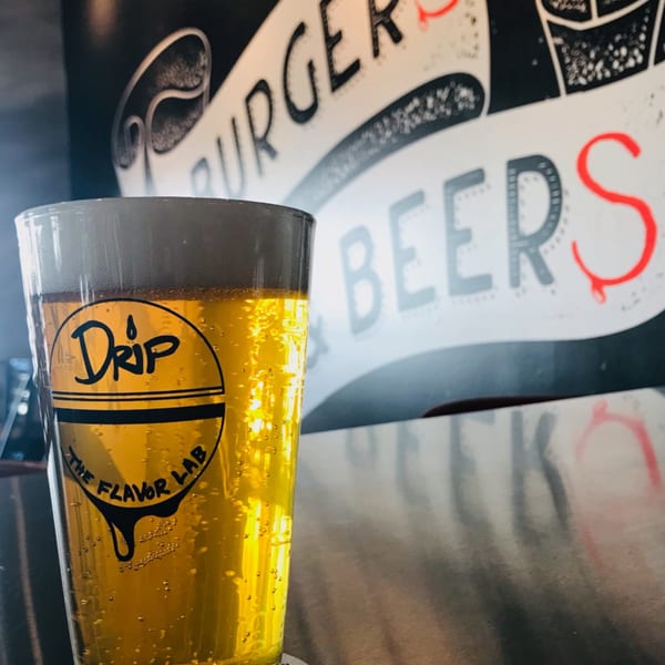 A Drip branded glass of golden beer with foam, against a wall mural reading "BURGERS BEERS".