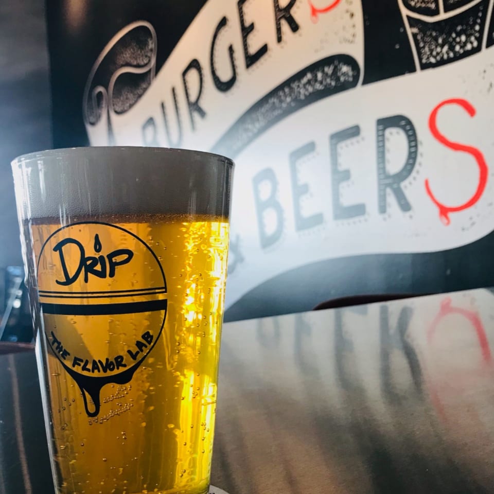 A Drip branded glass of golden beer with foam, against a wall mural reading "BURGERS BEERS".